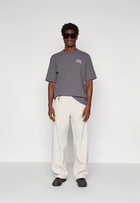 Hombre vistiendo una camiseta gris de gran tamaño con gráfico, pantalones blancos holgados, zapatos negros y grandes gafas de sol negras, de pie frente a un fondo blanco.