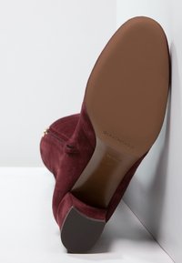Bota de tacón alto de ante burdeos con cremallera lateral, textura suave y punta redonda. Presenta una suela de goma marrón sólida.