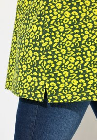 Grøn top med gul leopardprint, med lige kant og lette sidespalter, parret med mørkeblå jeans. Blød stoftekstur.