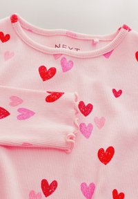 Pink ribbet langærmet top med farverige hjerte-mønstre i rød og pink. Har rund halsudskæring og bølgede manchetter. Brandlabel synlig.