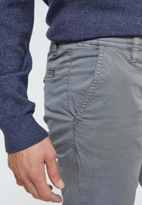Chinos en coton gris avec des détails de couture sur les poches et la ceinture, portés avec un pull en laine bleu marine, montrant une main reposant sur la hanche.