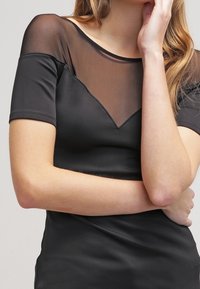 Černý top s průhledným síťovaným dekoltem, krátkými rukávy a přiléhavým střihem. Hladký materiál s trojúhelníkovým výřezem v oblasti hrudníku.