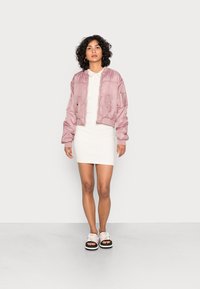 Casaco bomber rosa, vestido polo branco e sandálias chunky com sola preta. Cabelo encaracolado, em pé sobre um chão de cor clara contra um fundo simples.