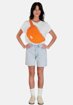 Femme debout, les mains sur les hanches, portant un t-shirt blanc, un short en jean clair, des baskets beiges, des chaussettes blanches, un collier coloré et un sac bandoulière orange vif.