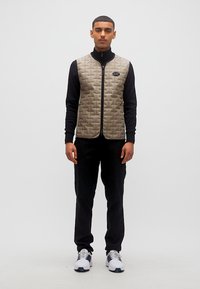 Άνδρας που φοράει μπεζ γιλέκο με καπιτονέ ύφασμα πάνω από μαύρο μακρυμάνικο πουκάμισο, μαύρο παντελόνι και λευκά αθλητικά παπούτσια, στέκεται μπροστά σε απλό φόντο.