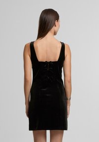 �Černé sametové šaty se čtvercovým výstřihem, zaobleným dekoltem a ozdobnými mašlemi. Hladká textura a přiléhavý střih.