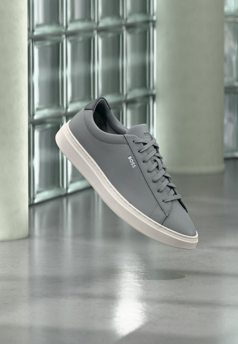 Baskets grises de la marque BOSS avec semelle blanche, lacées, flottant à l'intérieur devant un mur en blocs de verre et un sol en béton poli.