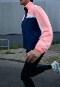 Chaqueta deportiva ligera en azul marino y rosa brillante con cremallera frontal, puños elásticos y diseño a bloques de color. Usada durante la carrera.
