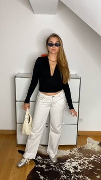 Blusa preta de mangas longas com decote em V profundo, jeans brancos de perna larga, ténis prateados e uma bolsa branca. Óculos de sol pretos complementam o visual.