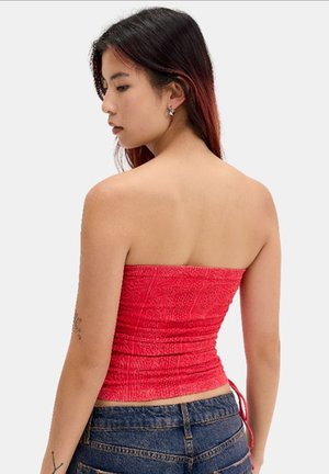 Femme portant un top rouge texturé sans bretelles et un jean bleu, de dos, regardant sur le côté sur un fond clair uni.