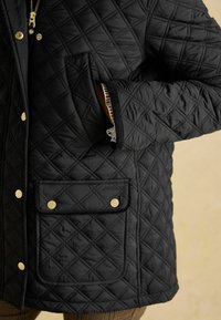 Jachetă neagră matlasată, cu modele în diamant, butoane aurii și un buzunar frontal. Material texturat cu margini contrastante pe mâneci.