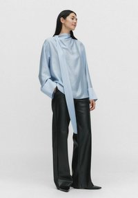 Vaaleansininen pitkähihaisessa puserossa on korkea kaulus ja edessä valuva solmu, yhdistettynä mustiin leveisiin tekonahkahousuihin.