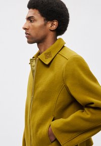 Volnen plašč gorčično rumene barve z zadrgo, strukturiranim ovratnikom in stranskimi žepi. Vezenih detajlov na ovratniku dodaja značilen pridih.