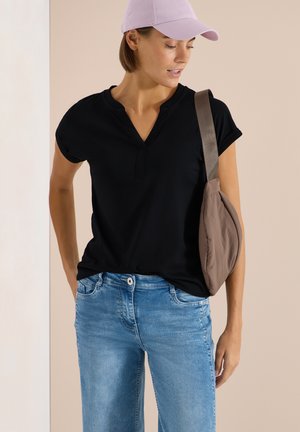 Donna con una maglietta nera a maniche corte, jeans chiari in denim, berretto lavanda e borsa a tracolla color taupe, che guarda verso il basso a sinistra.