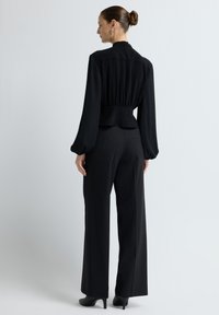 Sort bluse med samlet ryg, lange ærmer og høj krave, parret med sorte højtaljede bukser og hæle. Glat, letvægtsstof.