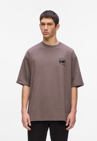 Jonge man in een oversized mauve t-shirt met een kleine zwarte grafiek van een gezicht met een zonnebril op de linkerborst, staand tegen een witte achtergrond.