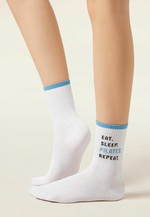 Coppia di calzini bianchi a coste con rifinitura blu, un calzino ricamato con "MANGIA. DORMI. PILATES. RIPETI." indossati ai piedi su sfondo semplice.