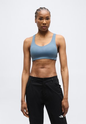 Urheilullinen nainen, jolla on sininen urheiluliivi ja mustat The North Face -housut, seisoo yksinkertaista vaaleaa taustaa vasten.