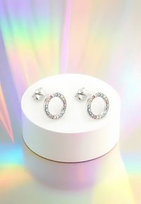 Silver runda studs med färgglada ädelstenar runt kanten, visade på en vit pedestal mot en holografisk bakgrund.