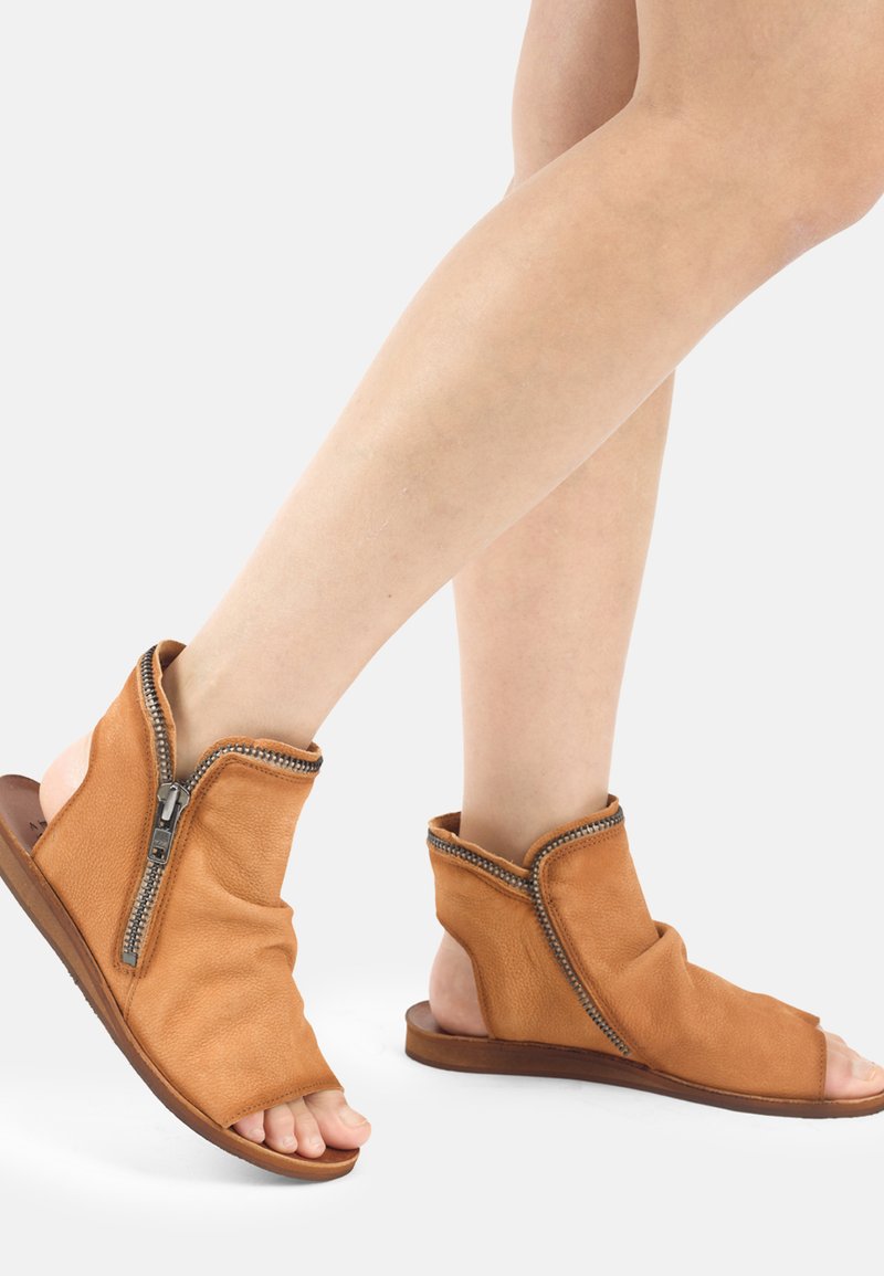 Jambes portant des sandales en cuir beige à bout ouvert, avec des fermetures éclair latérales visibles et des semelles plates basses, sur un fond clair uni.