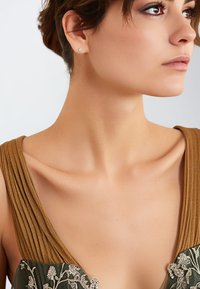 Braunes plissee Kleid mit tiefem V-Ausschnitt und grünen gestickten Akzenten. Das Model hat kurze Haare und trägt einen kleinen Stecker.