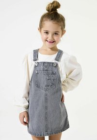 Grijze denim overall jurk met verstelbare banden, voorzakken en een licht uitlopende rok, gecombineerd met een witte blouse met pofmouwen.