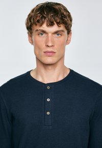 Maglietta henley a maniche lunghe di colore blu navy realizzata in tessuto testurizzato, con scollatura rotonda e tre bottoni marroni decorativi sul davanti.