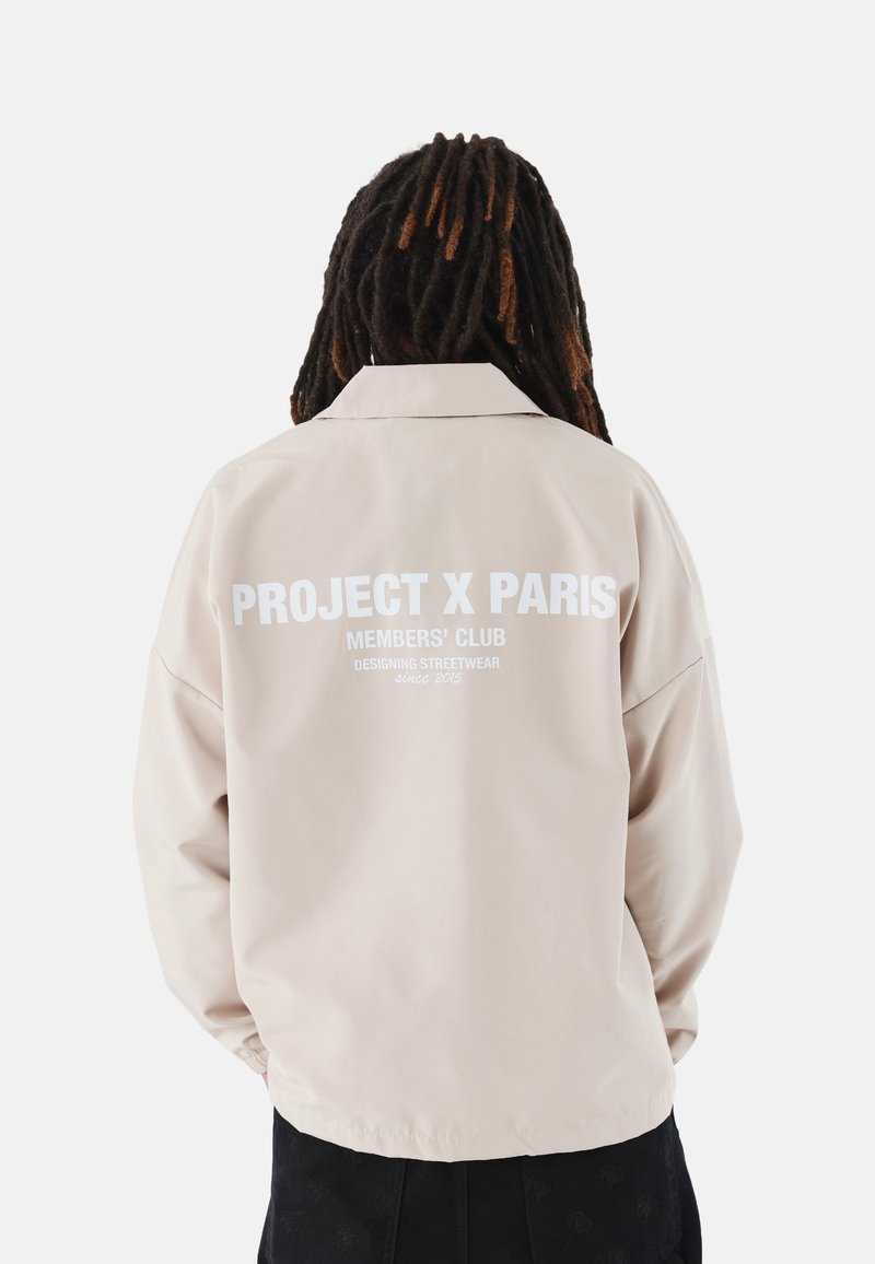 Chaqueta beige claro con un ajuste relajado, que presenta texto blanco en la parte trasera: "PROJECT X PARIS" y "MEMBERS' CLUB", fabricada con material suave.