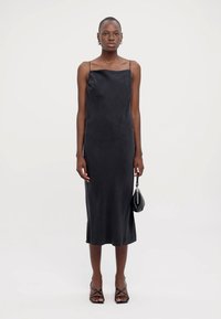 Black satin slip dress with thin straps, slim fit, mid-length, smooth texture, worn with a small black handbag and strappy sandals.