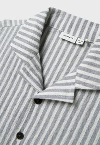 Camicia a righe bianco e grigia con una texture morbida, dotata di colletto a scollatura, bottoni neri e un'etichetta di abbigliamento.