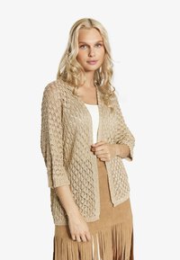 Stickad kofta i beige med ett texturerat, öppet mönster, 3/4-långa ärmar och en avslappnad passform, lagd över en vit topp och en fransad kjol.