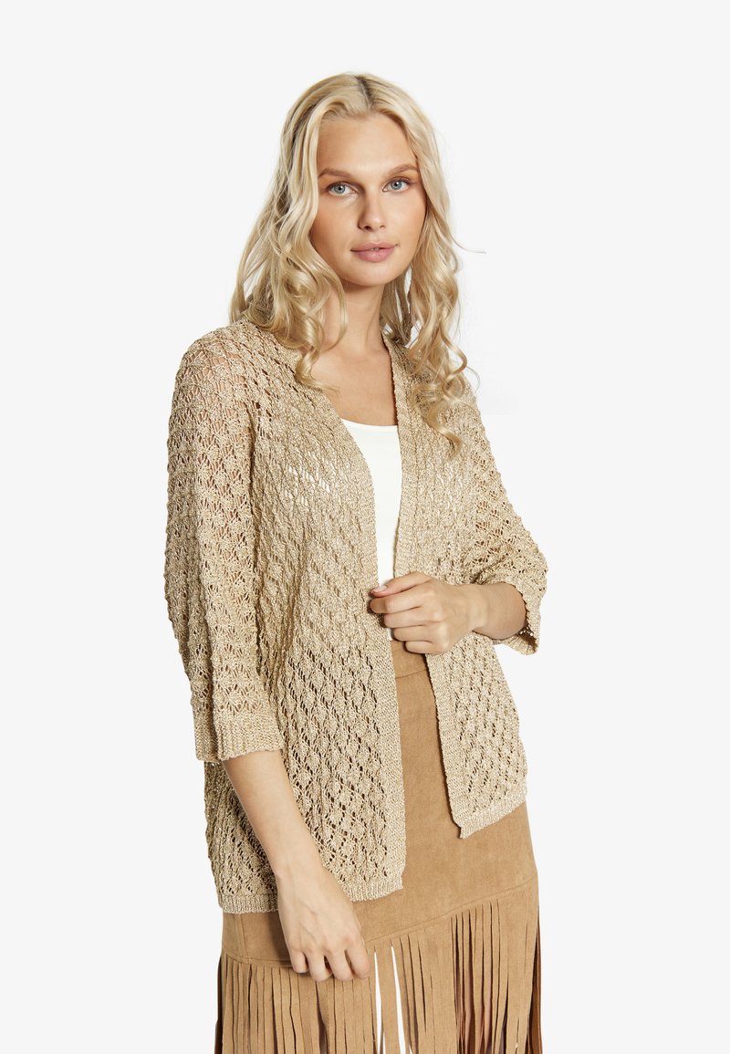 Stickad kofta i beige med ett texturerat, öppet mönster, 3/4-långa ärmar och en avslappnad passform, lagd över en vit topp och en fransad kjol.