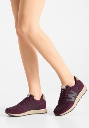 Benen die donkerpaarse mesh- en suède sneakers dragen met beige zolen en veters op een witte achtergrond.