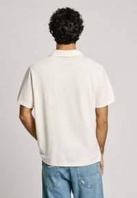 Short-sleeve, cream polo shirt with a classic collar, featuring a textured fabric and subtle stitching detail on the collar.