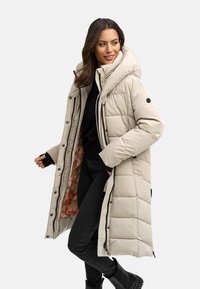 Beige langer Puffermantel mit hohem Kragen, Druckknöpfen und gestepptem Muster. Schwarze Innenschicht, getragen über einem schwarzen Pullover und einer schwarzen Hose.