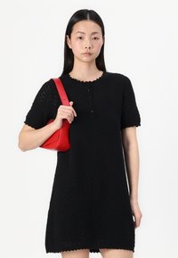 Črna pletena obleka z teksturiranim vzorcem, z valovitimi izrezi, kratkimi rokavi, v kombinaciji z majhno, strukturirano rdečo usnjeno torbico.