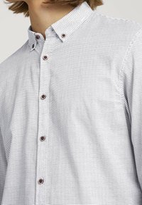 Biała koszula z długim rękawem w cienkie czarne pionowe paski. Posiada klasyczny kołnierzyk i brązowe guziki z przodu. Miękka tekstura materiału.