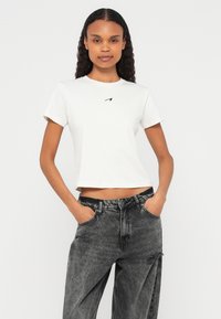 Νέα γυναίκα με σγουρά μαλλιά που φοράει λευκό κοντό T-shirt και ψηλόμεσα μαύρα τζιν με όξινο πλύσιμο, με τα χέρια στις τσέπες.