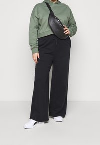 Sudadera verde recortada, pantalones de pierna ancha negros y riñonera negra. Zapatillas deportivas blancas con detalles negros. Tejidos suaves y corte relajado.