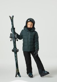 Dziecko w ciemnozielonej puchowej kurtce, czarnych spodniach narciarskich i rękawiczkach, trzymające czarne i niebieskie narty z wiązaniami, noszące czarny kask i gogle.