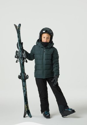 Bambino con giacca imbottita verde scuro, pantaloni da sci neri e guanti, che tiene sci neri e blu con attacchi, indossa un casco nero e occhiali da sci.