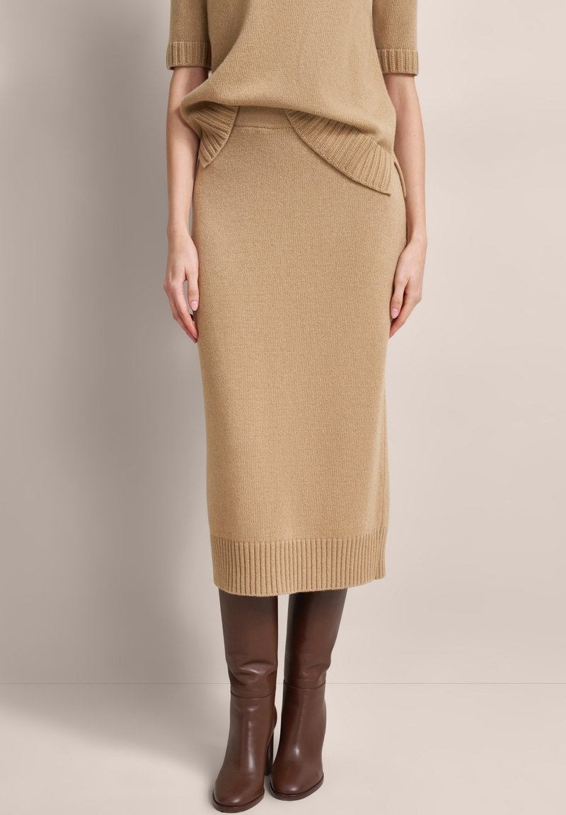 Beige Strickpullover und passender Midi-Rock mit geripptem Saum und Taschen, kombiniert mit knielangen braunen Lederstiefeln.