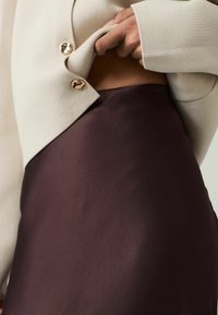 Blazer beige texturizzato con bottoni dorati, indossato sopra una gonna di satin bordeaux, che mette in risalto il tessuto morbido e una silhouette elegante.