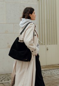 Black suede hobo bag with a slouchy design, featuring a smooth leather strap and metallic hardware accents. Model wears a beige trench coat.