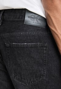 Schwarze Jeans aus Denim haben eine strukturierte Oberfläche, eine Gesäßtasche und ein rechteckiges schwarzes Lederetikett mit der Prägung "Calvin Klein Jeans".