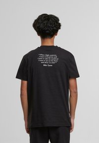 Černé bavlněné tričko s krátkým rukávem a kulatým výstřihem, které na zadní straně obsahuje bílý text s citátem přisuzovaným Mikeu Tysonovi.