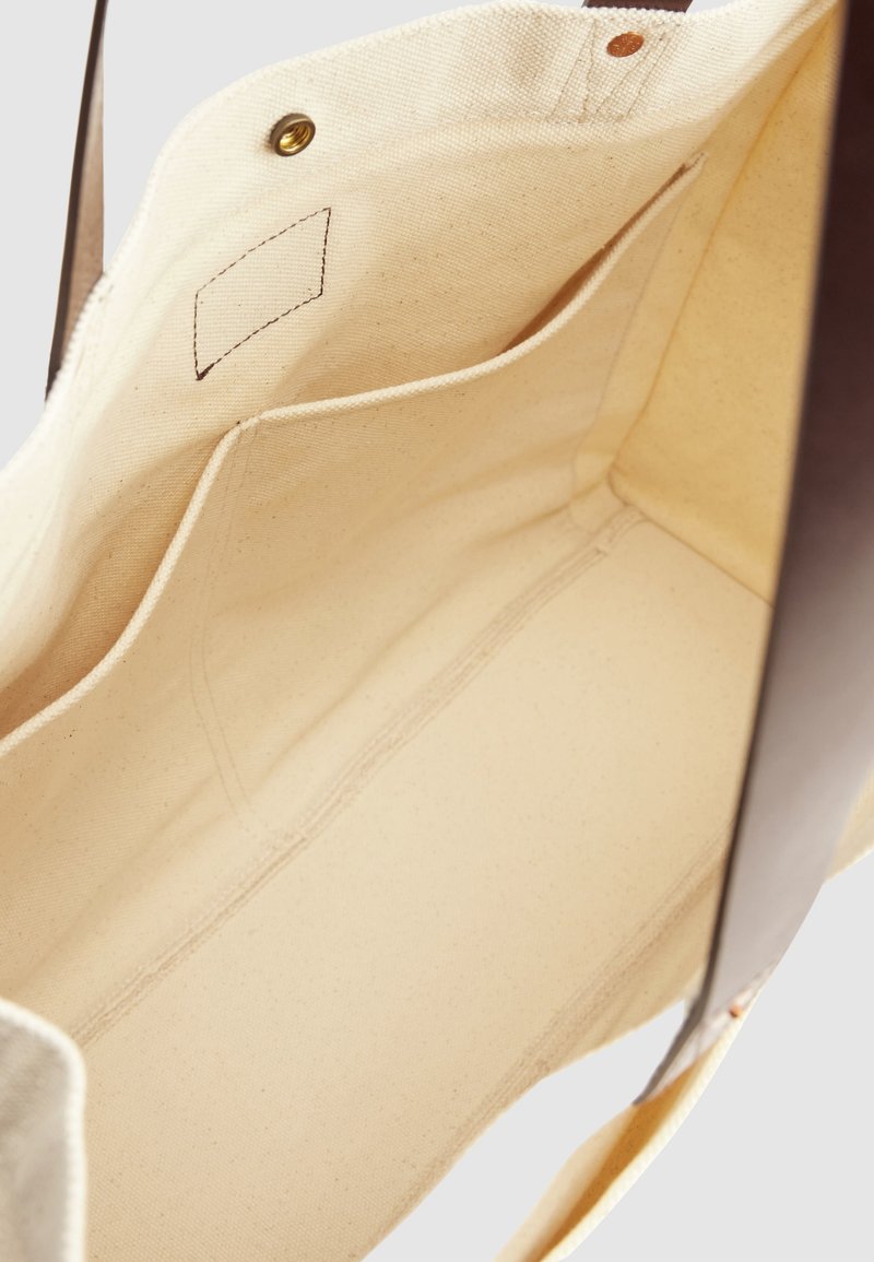 Innenraum einer beigen Canvas-Tragetasche mit einer offenen Tasche und braunen Ledergriffen, gesichert durch einen Messing-Druckknopf.