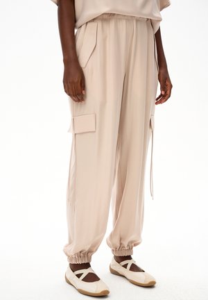 Beżowe spodnie typu cargo z elastycznymi ściągaczami noszone z beżowymi balerinami z krzyżującymi się paskami na ciemnej skórze.