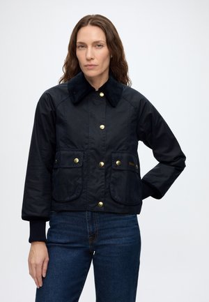 Femme aux longs cheveux bruns portant une veste noire avec des boutons dorés et un jean bleu, debout avec une main sur la hanche.