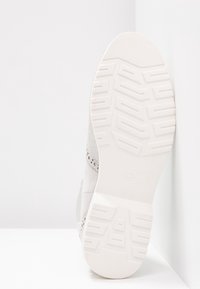 Semelle d'une chaussure de couleur claire avec une semelle à motif, positionnée debout contre un mur blanc et une surface blanche.